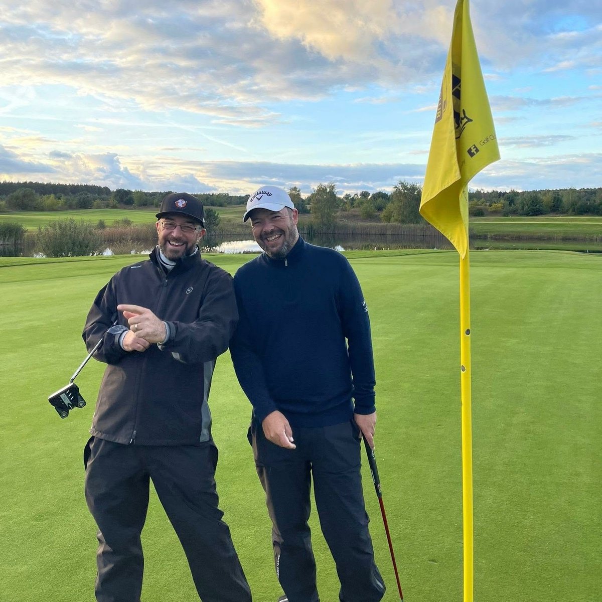 Two golfers posing together on a green golf course with a yellow flagstick, wearing dark clothing and golf caps on a sunny day.