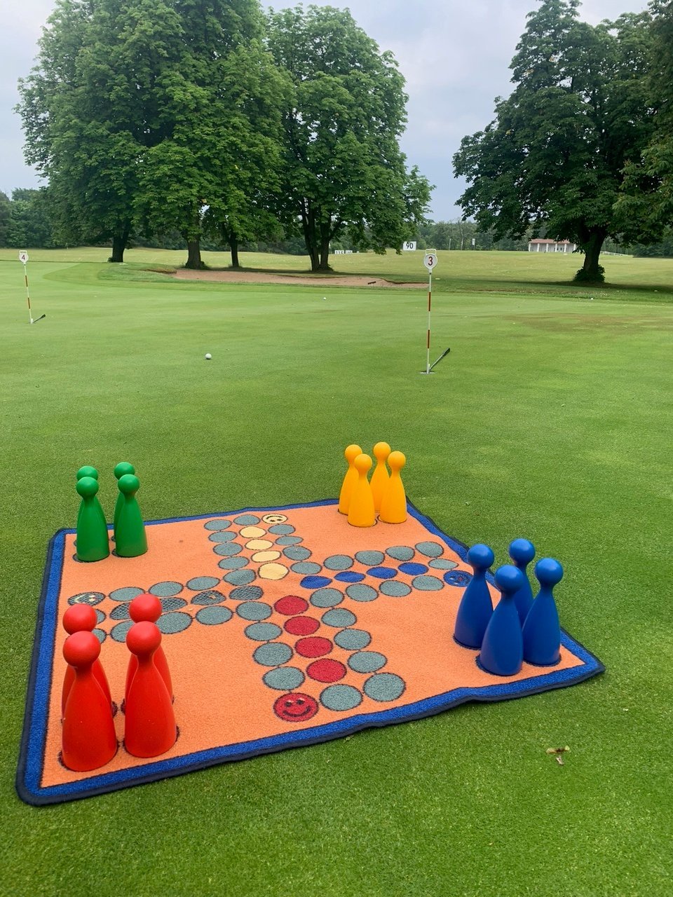 Oversized checkers game board set up on grass with red, yellow, blue, and green game pieces in a park setting