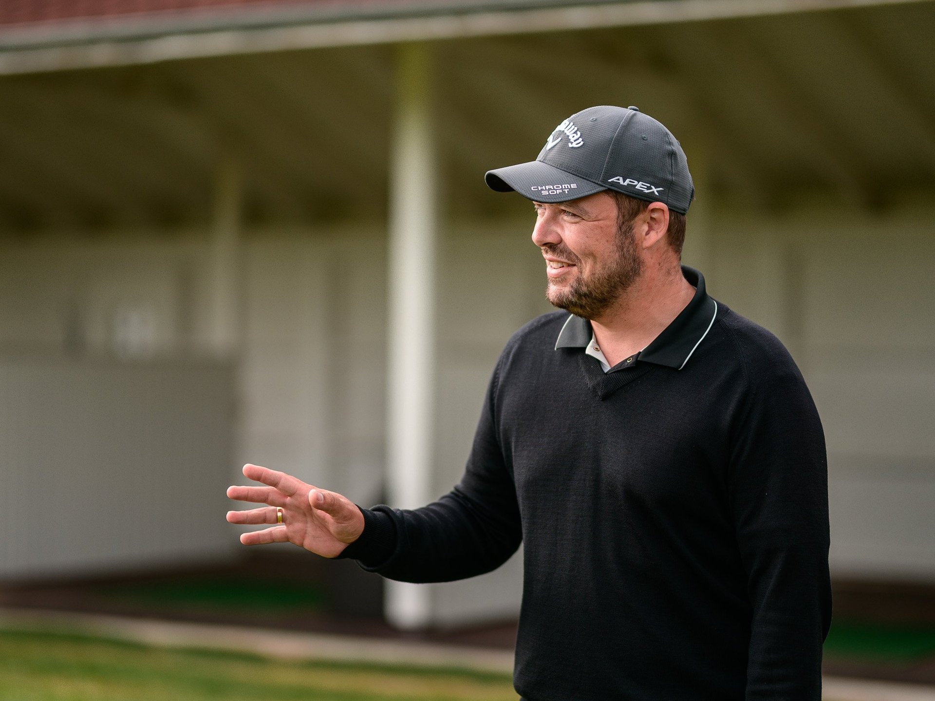 Man in black polo shirt and cap gesturing while speaking outdoors near a garage